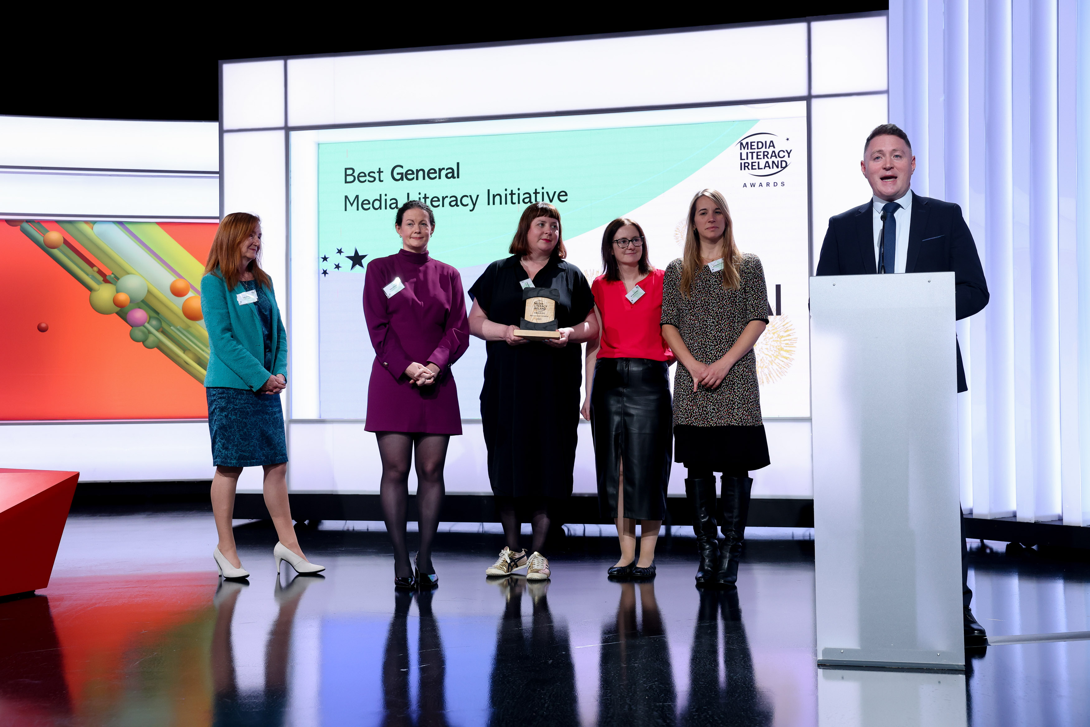 Five women standing in front of a screen which reads 'Best General Media Literacy Iniative'. There is a man speaking at a podium to the right.